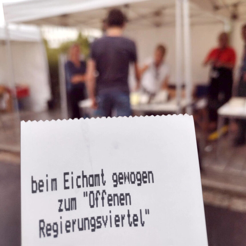 Im Vordergrund sieht mal einen Zettel mit der Aufschrift: beim Eichamt gewogen zum "Offenen Regierungsviertel". Im Hintergrund sieht man eine Person, die gerade auf die Personenwaage steigt. Das Foto entstand beim Tag des offenen Regierungsviertel 2024.