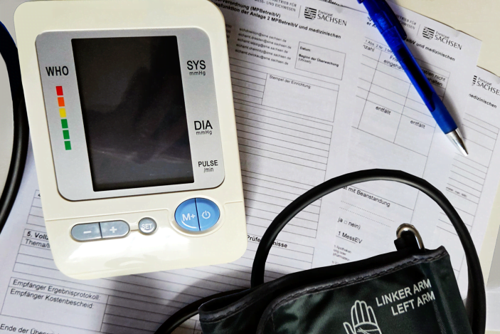 Blutdruckmessgerät mit Manschette für den linken Arm und Anzeigegerät liegt auf Ausdrucken von Protokollvorlagen der sächsischen Eichbehörde für den medizinischen Bereich