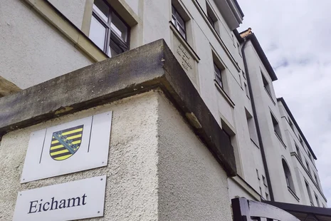 Schild mit Sachsen-Wappen und darunter der Aufschrift „Eichamt“. Im Hintergrund sieht man das helle Gebäude des Eichamtes Chemnitz.