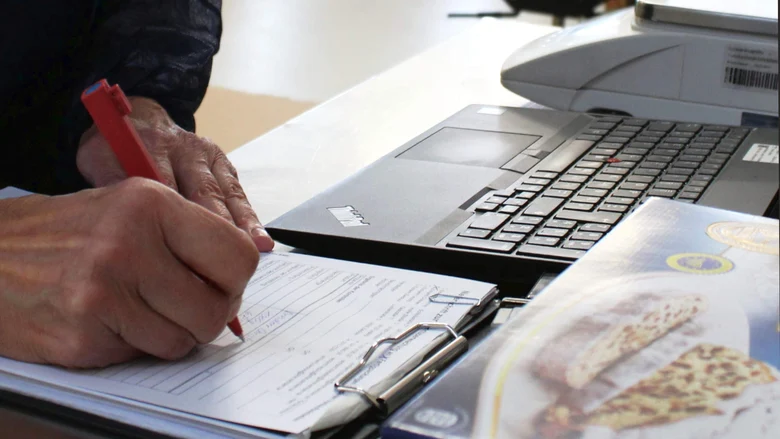 Im Vordergrund sieht man die Hände einer Person, die Daten in ein Prüfprotokoll schreiben. Daneben steht eine Fertigpackung mit Weihnachtsstollen. Im Hintergrund sieht man einen Laptop und eine Kontrollwaage.