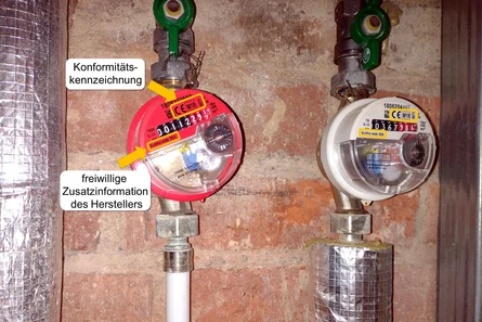 Zwei Wasserzähler an einer Ziegelwand. Ein gelber Pfeil markiert die Konformitätskennzeichnung, ein weiterer zeigt auf die freiwillige Zusatzinformation des Herstellers „Eichfrist endet 2024“.