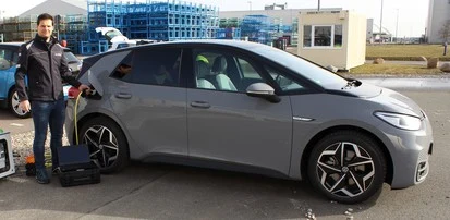 Ein Mann steht an einem grauen Elektrofahrzeug. Er steckt gerade den Ladestecker ein. Das Fahrzeug steht auf einem großen Parkplatz, im Hintergrund sieht man ein blau eingezäuntes Gebäude.