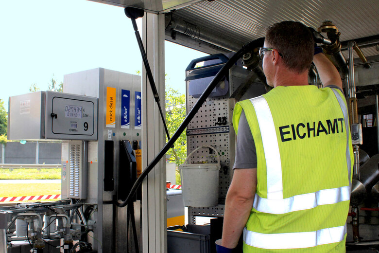Ein Mann steht mit dem Rücken zur Kamera. Auf seiner Warnweste steht „Eichamt“. Er hält eine Zapfpistole in einen Kolben, der in seinem Prüffahrzeug verbaut ist und schaut zur Anzeige an der Zapfsäule. Die Anzeige lautet: 274,34 €, 154,21 Liter.