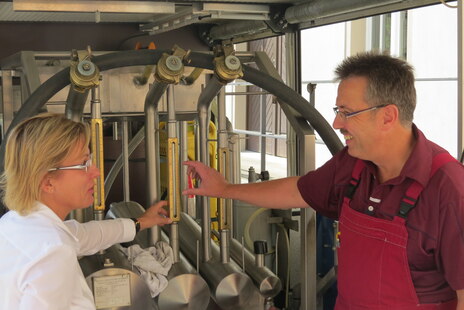 Ein Kollege vom Eichamt steht mit Frau Klepsch (damals Ministerin für Soziales und Verbraucherschutz) vor Prüfkolben. Beide lächeln. Er deutet mit Daumen und Zeigefinger eine Spanne an und zeigt damit auf die Skalierung am Prüfkolben.