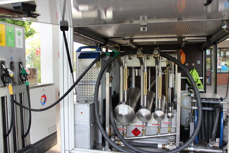 Prüffahrzeug zur Eichung von Zapfsäulen an der Tankstelle. Darin sind vier unterschiedlich große Kolben verbaut, an deren Einflussrohr eine Skalierung zu erkennen ist. Eine Zapfpistole steckt im größten Kolben. Neben dem Fahrzeug sieht am drei Zapfpunkte.