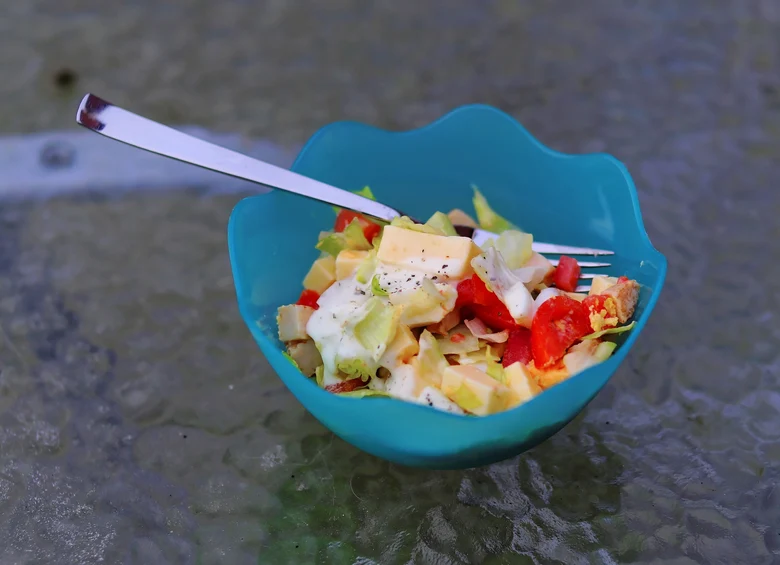 Blaue Dessertschüssel auf einer Glaspaltte. In der Schüssel ist ein gemischter Salat mit Eisbergblättern, Tomate, Gurke, Käse und Joghurtdressing. In dem Salat steckt eine Gabel.